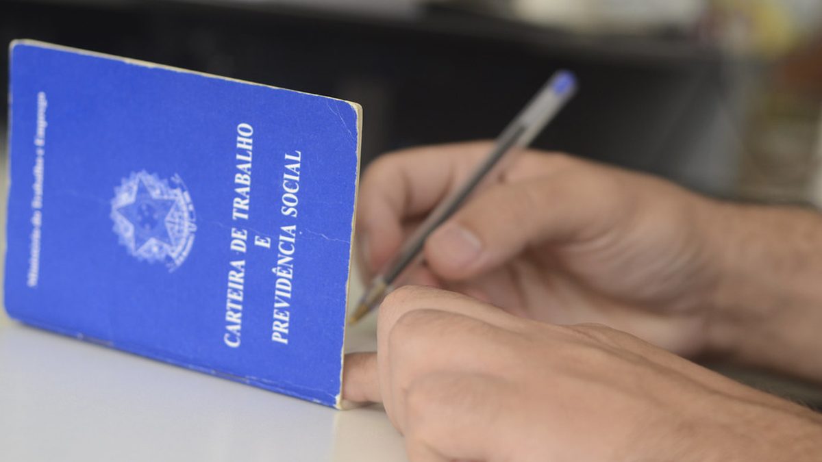 Carteira de trabalho azul sobre a mesa, ao lado de mãos que assinam um papel